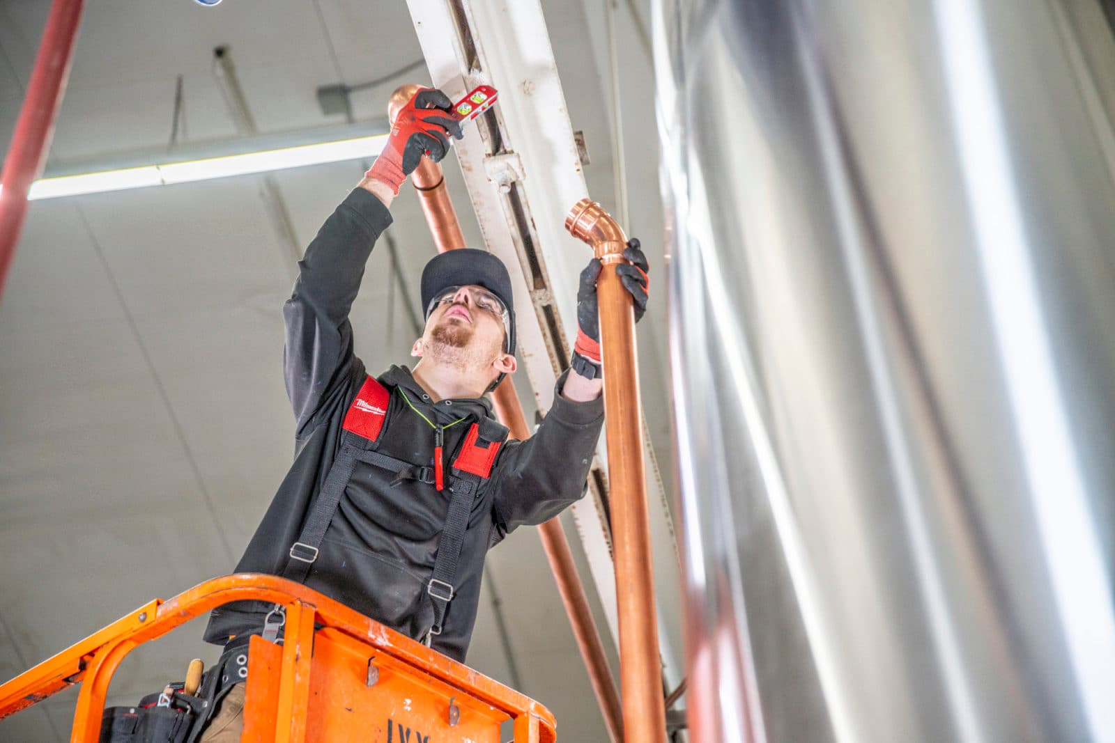 Low-angle plumber on lift beside stainless tanks